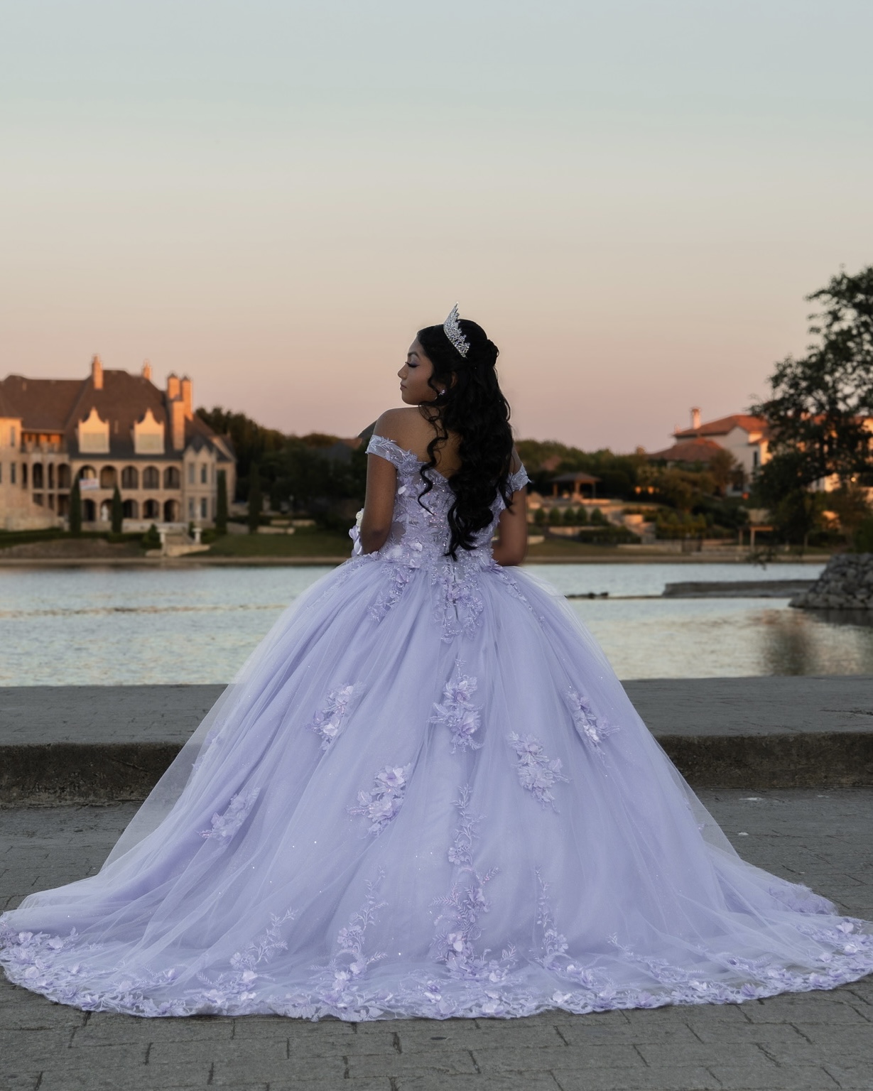 A quinceañera in a lavender gown and tiara posing by the water at sunset